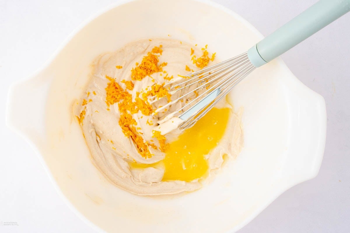 A mixing bowl with pale batter, orange zest, and melted butter being combined with a whisk on a white surface.
