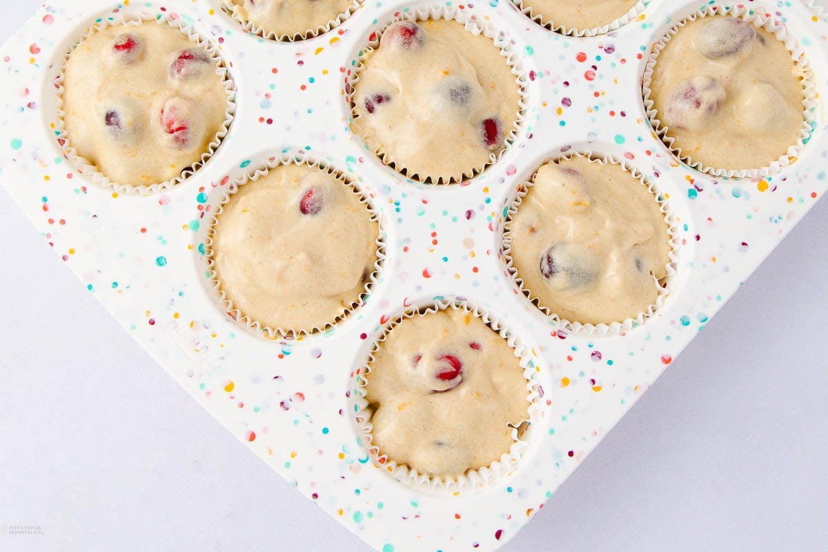 A close-up of a muffin tin lined with cupcake papers, filled with unbaked batter containing visible cranberries or berries, ready to be baked. The muffin tin has a colorful confetti pattern.