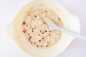 A white mixing bowl containing thick batter with cranberries and a gray spatula resting inside, all on a white surface.