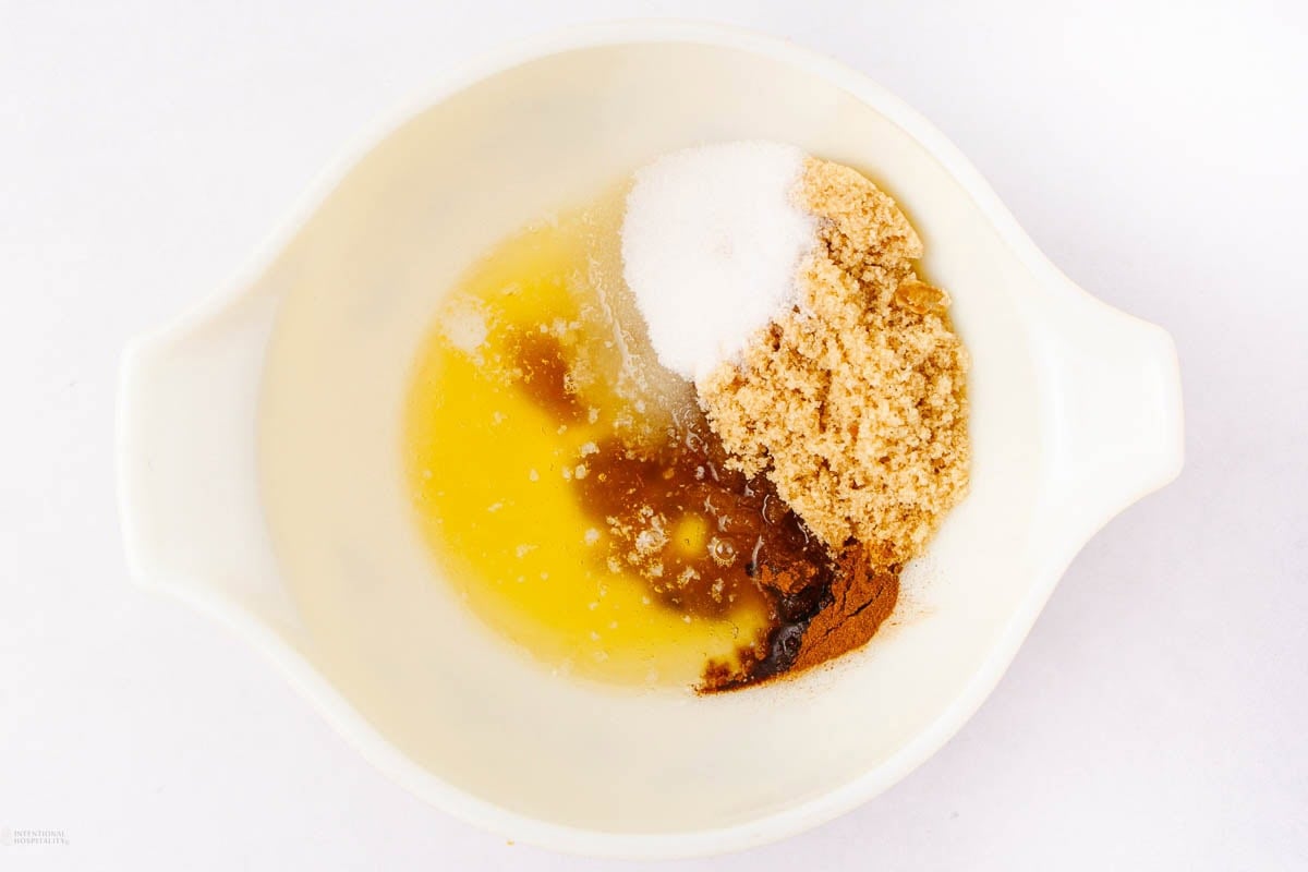 A white mixing bowl containing melted butter, granulated sugar, brown sugar, cinnamon, and vanilla extract, all separated into sections on a white background.