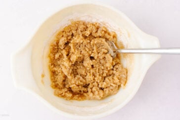 A mixing bowl containing thick, light brown cookie dough with a metal spoon resting in it, set on a white surface.