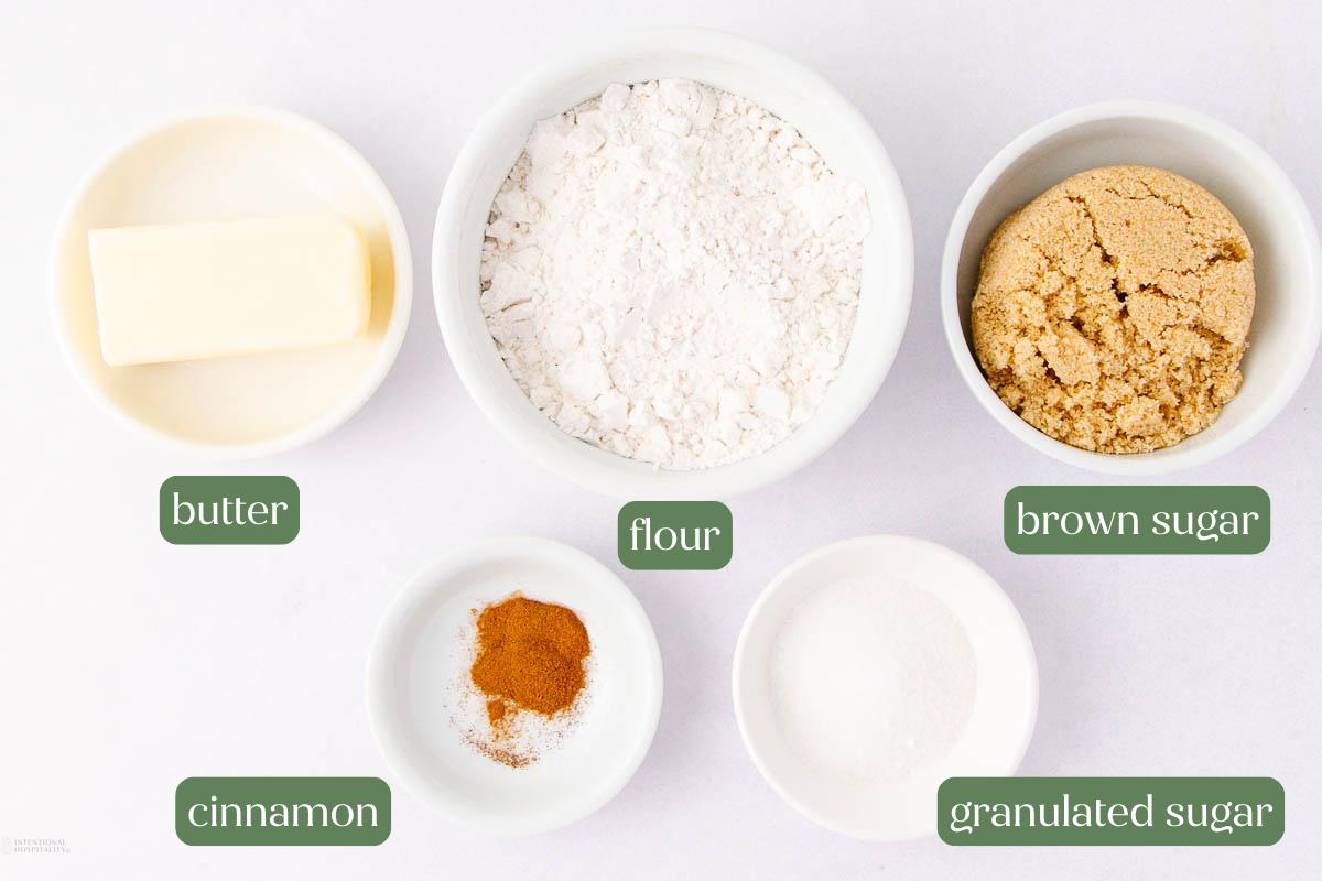 Five white bowls on a white surface containing butter, flour, brown sugar, cinnamon, and granulated sugar, each labeled with green text.