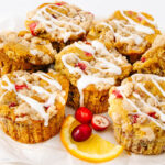Seven cranberry-orange muffins with crumb topping and white icing drizzle, arranged on parchment paper with fresh cranberries and an orange slice for garnish.