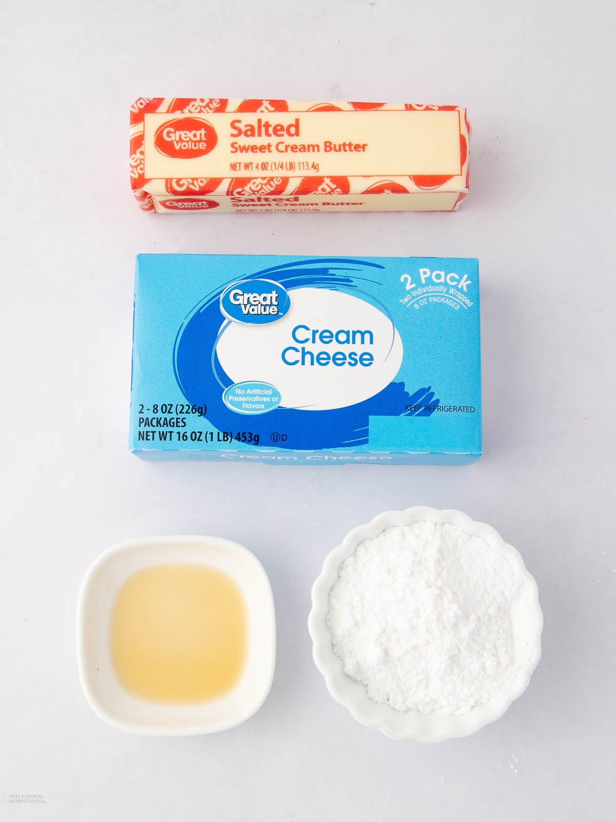 A stick of salted butter, a package of cream cheese, a small bowl of vanilla extract, and a small bowl of powdered sugar on a white surface.