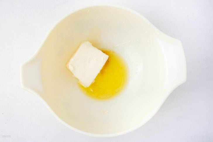 A white mixing bowl contains a chunk of butter and some melted butter, set against a plain white background.