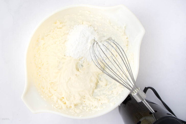 A white mixing bowl with creamy batter and a mound of powdered sugar, being mixed with a metal whisk attached to an electric hand mixer, on a white surface.