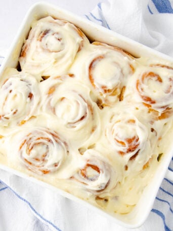 A white baking dish filled with seven frosted cinnamon rolls covered in creamy icing, resting on a white surface with a blue and white striped cloth nearby.