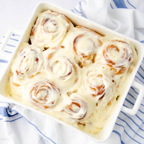 A white baking dish filled with seven frosted cinnamon rolls covered in creamy icing, resting on a white surface with a blue and white striped cloth nearby.