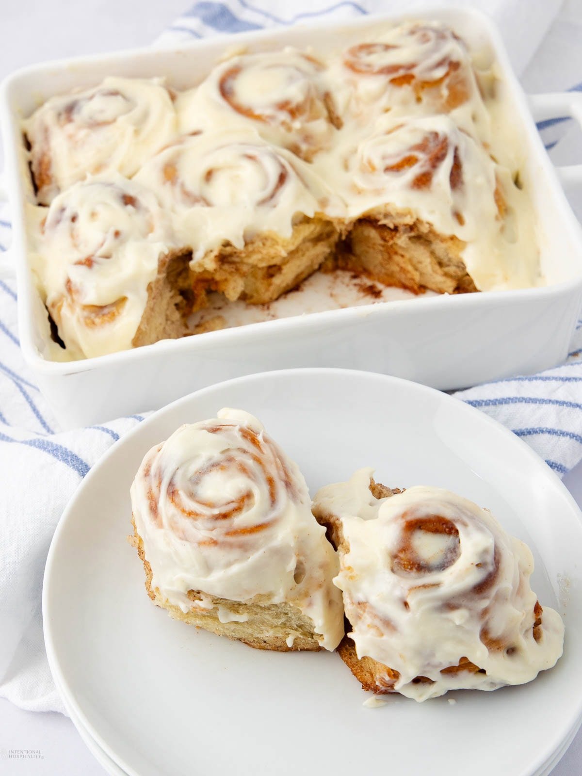 A plate with two frosted cinnamon rolls sits in front of a baking dish filled with more cinnamon rolls, all topped with creamy white icing. The background features a white and blue striped cloth.