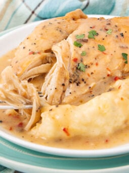 A plate of shredded chicken breast with gravy, served alongside a generous portion of mashed potatoes, garnished with chopped herbs. A fork is pulling apart the tender chicken. A plaid cloth is in the background.