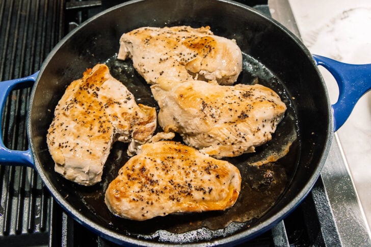 Four seasoned chicken breasts are being cooked in a black cast iron skillet on a stovetop, with the chicken pieces showing golden brown seared surfaces.