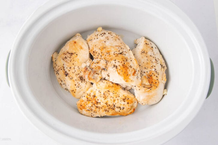 Four cooked, seasoned chicken breasts arranged in a white slow cooker on a white surface.