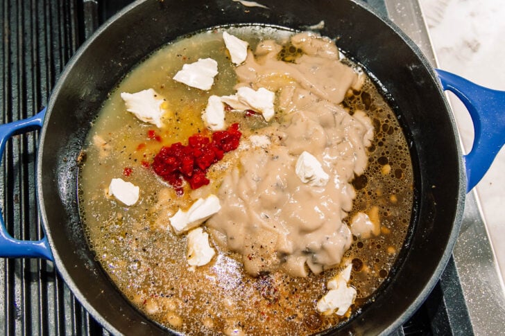 A blue cast iron pot on a stove contains a mixture of cream soup, red berries or cranberry sauce, small dollops of cream cheese, broth, and seasonings, all about to be stirred together.