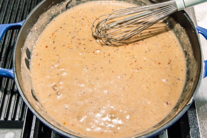 A whisk stirring creamy, pepper-speckled gravy in a blue skillet on a stove.