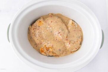 Chicken breasts covered in a creamy, speckled sauce sit inside a white slow cooker, ready to be cooked. The sauce appears thick and contains visible herbs and spices.