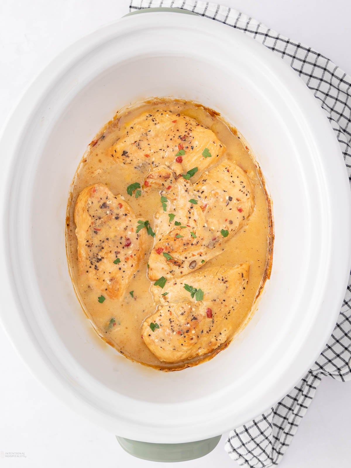 Four cooked chicken breasts in a creamy, seasoned sauce, garnished with parsley, inside a white slow cooker. A black-and-white checkered cloth is placed underneath the cooker.