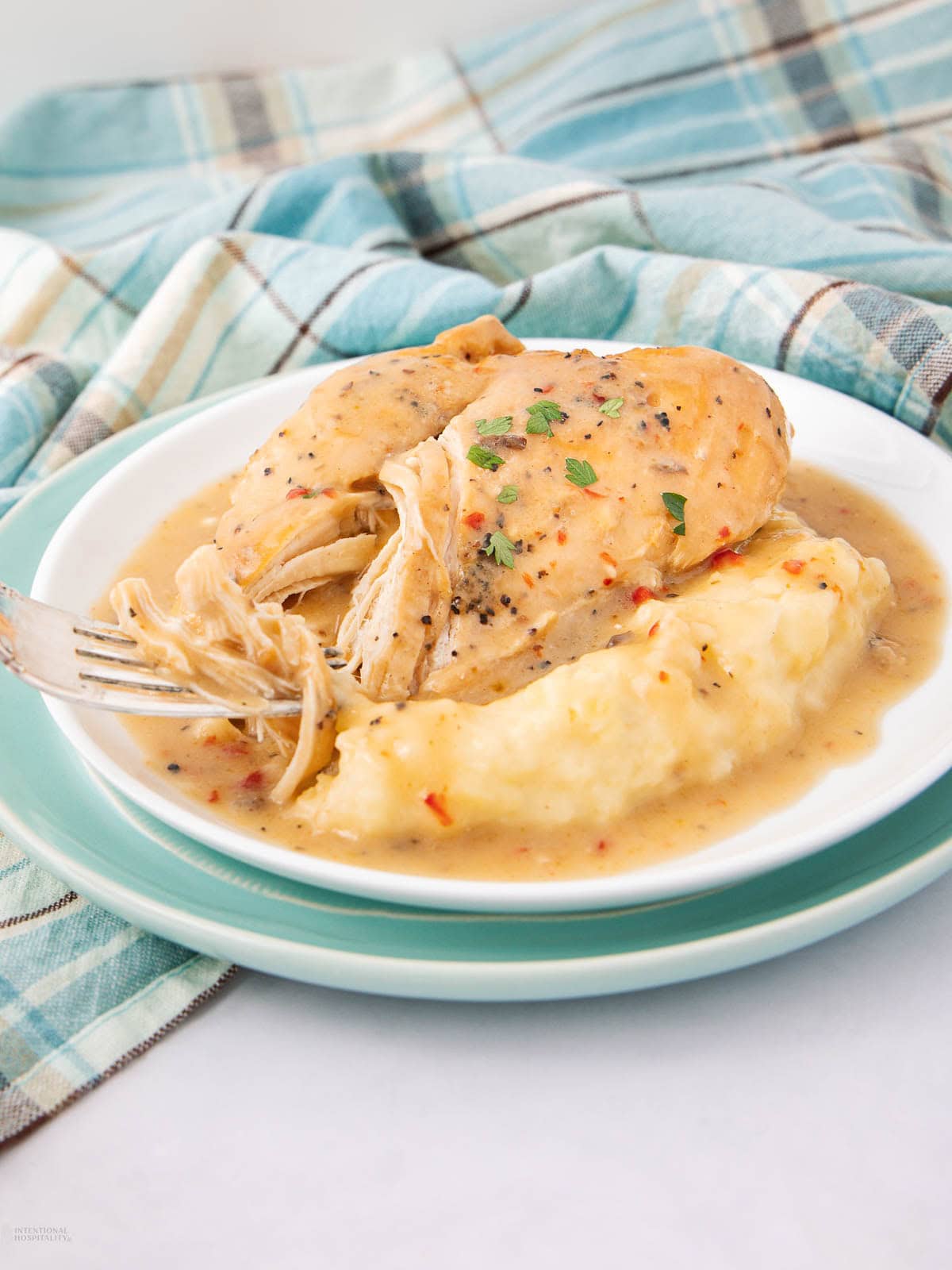 A plate with shredded chicken breast, creamy mashed potatoes, and gravy, garnished with herbs, sits on a teal and white dish with a blue plaid cloth in the background.