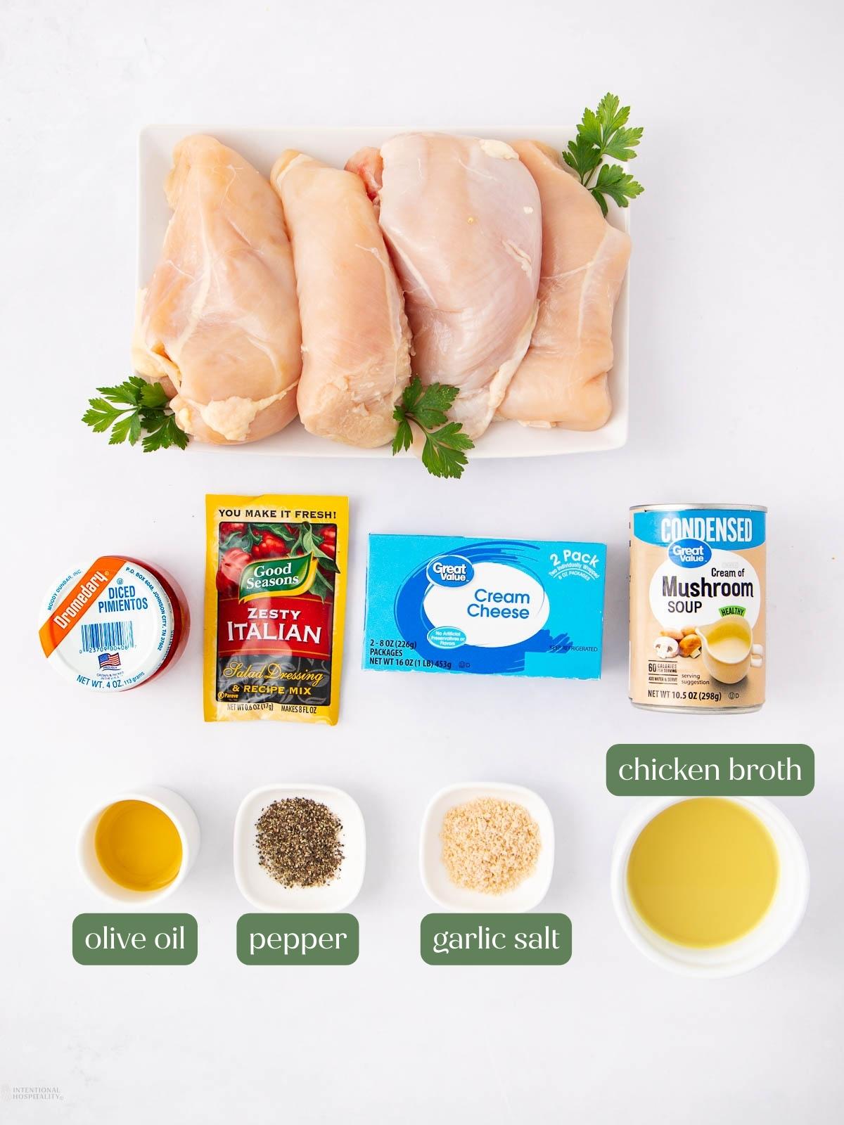 Overhead view of ingredients for a chicken recipe: raw chicken breasts on a plate with parsley, diced pimentos, Italian seasoning, cream cheese, cream of mushroom soup, olive oil, pepper, garlic salt, and chicken broth in bowls.