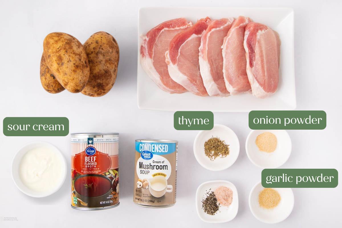 Top-down view of raw pork chops, whole potatoes, sour cream, canned beef broth, canned cream of mushroom soup, dried thyme, onion powder, and garlic powder arranged on a white surface.