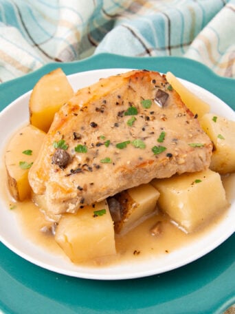 A cooked pork chop with gravy sits atop chunky pieces of potatoes and turnips on a white plate, garnished with chopped herbs. The plate is on a teal dish, with a plaid cloth in the background.