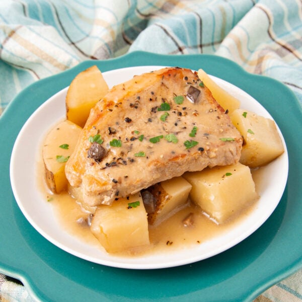 A cooked pork chop with gravy sits atop chunky pieces of potatoes and turnips on a white plate, garnished with chopped herbs. The plate is on a teal dish, with a plaid cloth in the background.