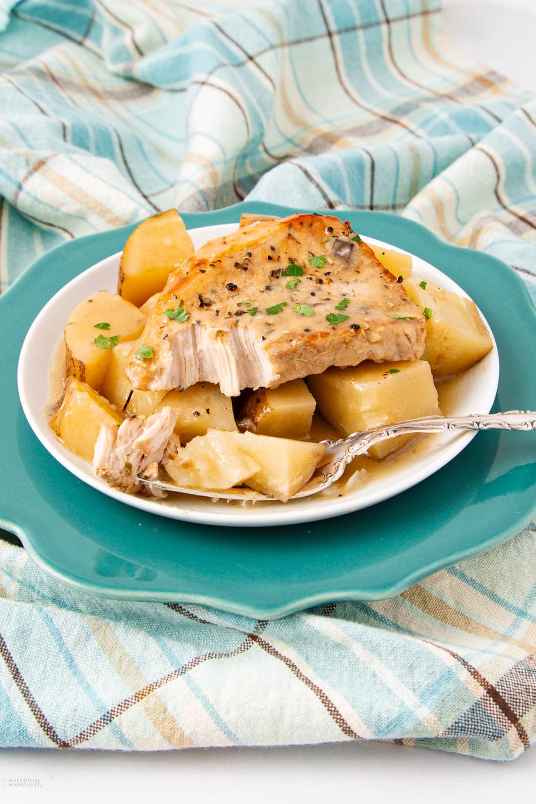 A plate with a pork chop and chunks of potatoes, garnished with herbs, sits on a turquoise charger. A fork rests on the plate, and the background features a striped cloth in shades of teal and beige.