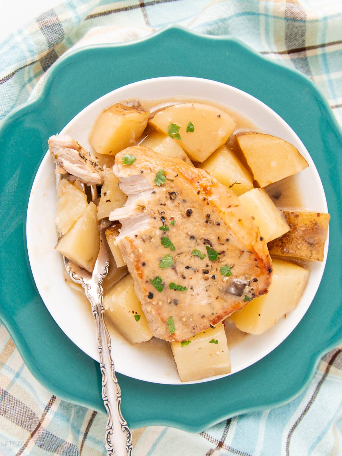 A plate with a cooked pork chop and chunks of potatoes garnished with herbs, served on a white bowl with a silver fork, placed on a teal plate over a plaid cloth.