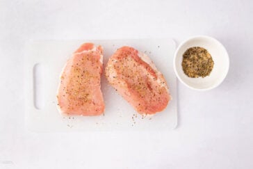 Two raw pork chops seasoned with herbs and spices sit on a white cutting board next to a small white bowl filled with additional dry seasoning, all on a white surface.