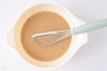 A light-colored mixing bowl filled with tan batter or sauce, with a light blue whisk resting inside on a white surface.