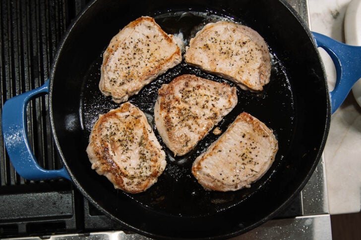 Five seasoned pork chops cooking in a black cast iron skillet with a blue handle, sitting on a stovetop. The pork chops are seared and arranged in a circular pattern.