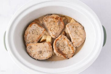 Five seasoned pork chops in creamy sauce sit atop sliced potatoes inside a white slow cooker, ready to be cooked.