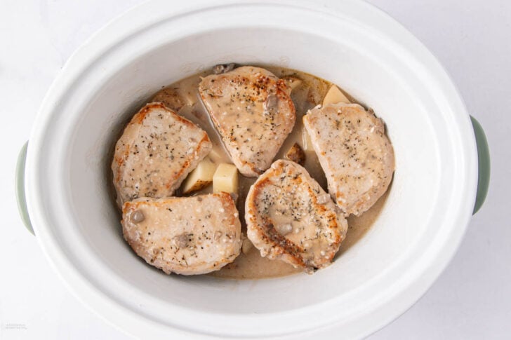 Five seasoned pork chops in creamy sauce sit atop sliced potatoes inside a white slow cooker, ready to be cooked.