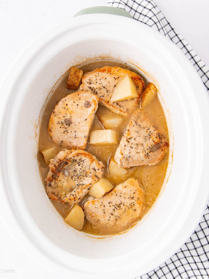 Four seasoned pork chops and potato chunks simmer in a light brown sauce inside a white slow cooker, placed on a black and white checkered cloth against a white background.