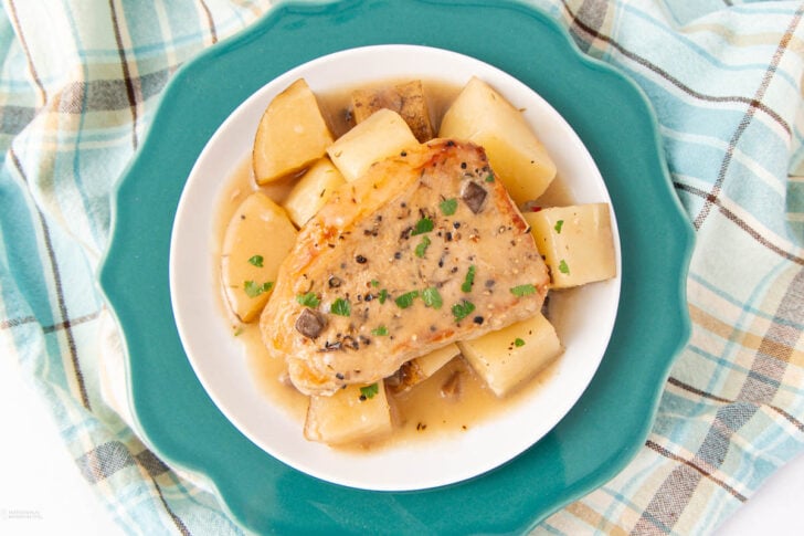 A plate with a seasoned pork chop in gravy, garnished with herbs, surrounded by chunks of cooked potatoes. The plate sits on a teal charger over a blue and beige plaid cloth.