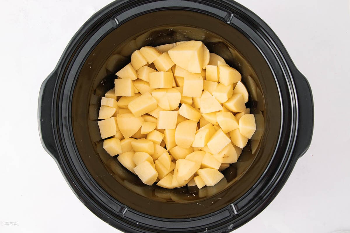 Chopped raw potatoes are placed inside a black slow cooker, ready for cooking. The slow cooker is viewed from above against a white background.