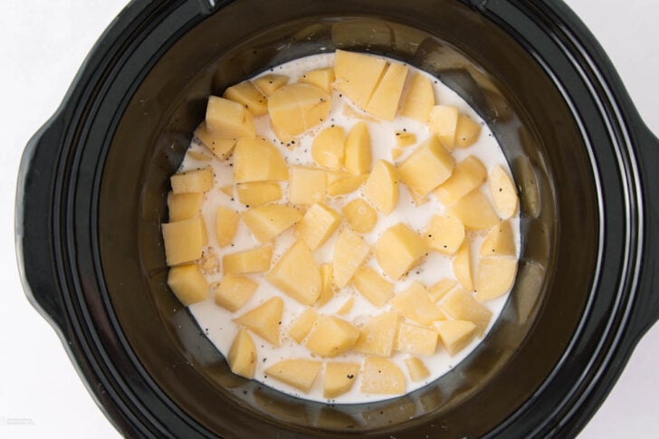 Diced potatoes in a slow cooker with milk and black pepper, ready to be cooked.