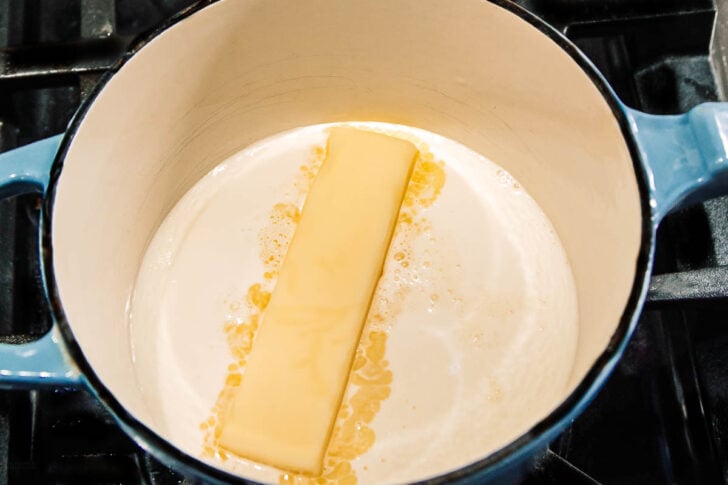 A stick of butter melting in the center of a white enameled pot on a stovetop.