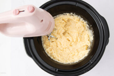 A pink electric hand mixer is blending mashed potatoes in a black slow cooker on a white surface.