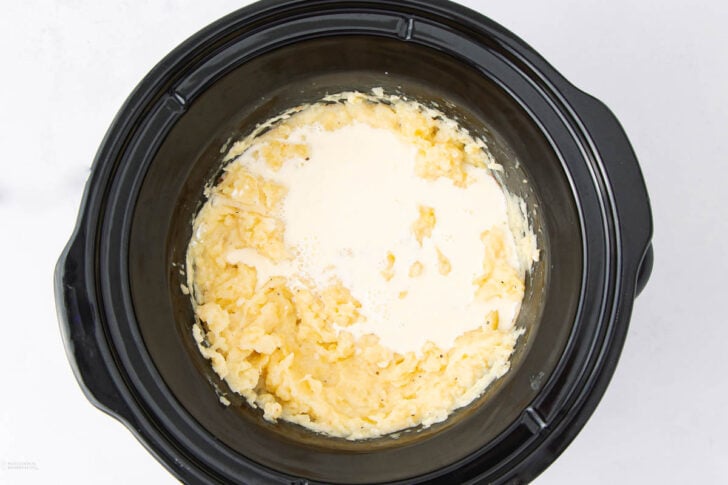A black slow cooker filled with partially mashed potatoes and cream on top, ready to be mixed together, sitting on a white surface.