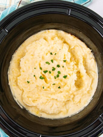 Creamy mashed potatoes garnished with chopped parsley in a black slow cooker, with a plaid cloth nearby.