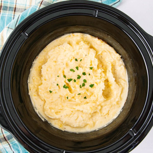 Creamy mashed potatoes garnished with chopped parsley in a black slow cooker, with a plaid cloth nearby.
