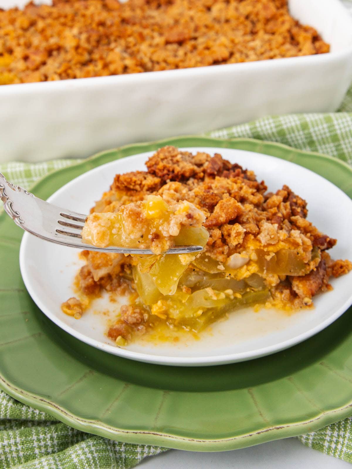 A close-up of a slice of apple crisp with a crumbly topping on a white plate, with a fork lifting a bite. A green napkin and the rest of the dessert in a baking dish are in the background.