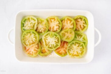 Sliced green tomatoes arranged in a white baking dish, seasoned with salt, pepper, and spices, ready for cooking.
