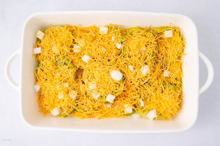 A white baking dish filled with sliced green bell peppers, topped with shredded cheddar cheese and small chunks of white onion, ready to be baked.