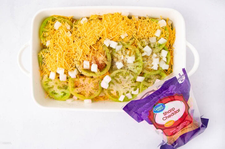 A white baking dish filled with sliced green tomatoes, chopped onions, shredded sharp cheddar cheese, and seasonings, with an open bag of sharp cheddar cheese in the foreground.
