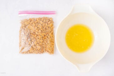 A plastic bag of crushed graham crackers sits next to a white mixing bowl filled with melted butter on a white surface.