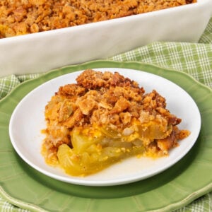 A slice of green tomato crisp with a golden, crumbly topping sits on a white plate with a green rim. A baking dish with more crisp is in the background on a green plaid cloth.