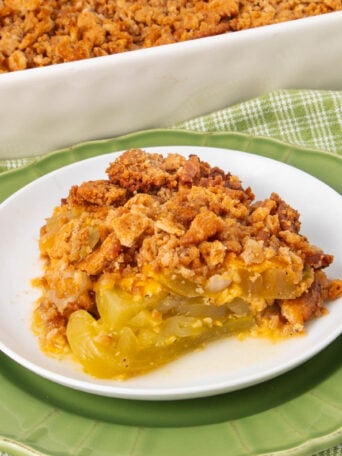 A slice of green tomato crisp with a golden, crumbly topping sits on a white plate with a green rim. A baking dish with more crisp is in the background on a green plaid cloth.