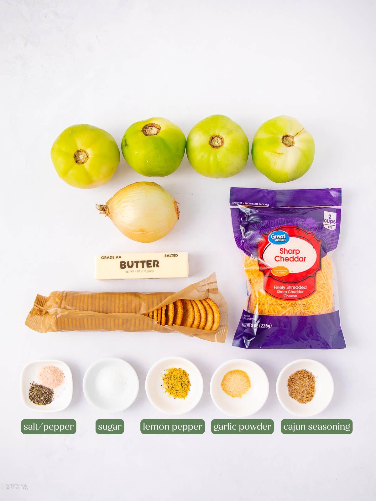 Overhead view of ingredients for a recipe: three green tomatoes, an onion, a stick of butter, sharp cheddar cheese, a sleeve of crackers, and small bowls of salt, pepper, sugar, lemon pepper, garlic powder, and Cajun seasoning.