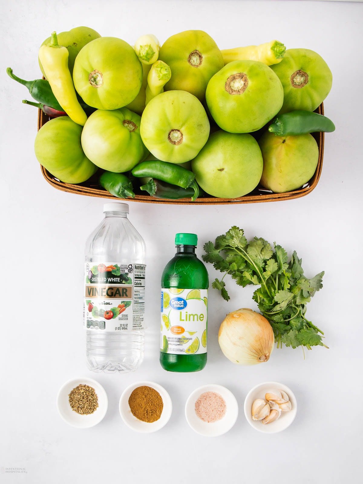 A basket of green tomatoes, peppers, and tomatillos sits above bottles of white vinegar and lime juice, fresh cilantro, an onion, four garlic cloves, and small bowls of dried spices and pink salt on a white surface.